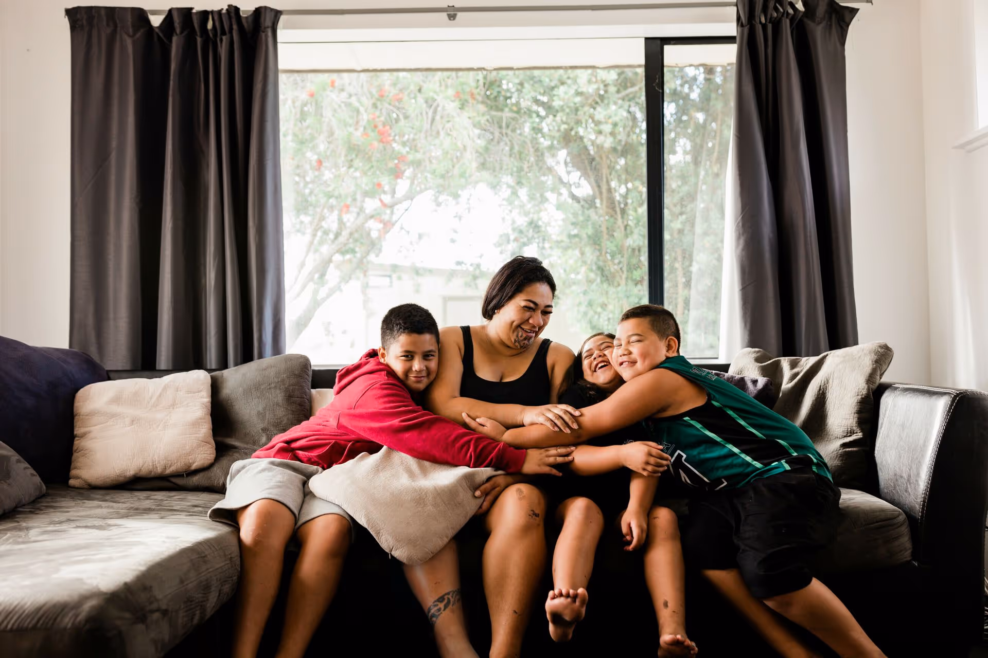 A woman sits on a couch, surrounded by three children hugging her, with sunlight filtering through a window in the background.