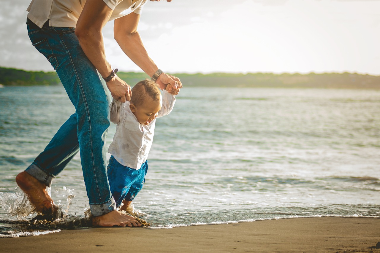 Two young children hold hands in shallow water at the beach, smiling and playing together as the sun sets behind them.