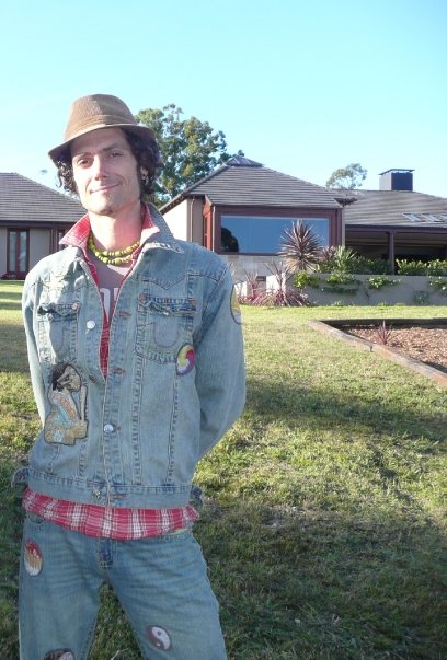 Man wearing a brown hat, denim jacket with patches, and plaid shirt standing on grass in front of a modern house.