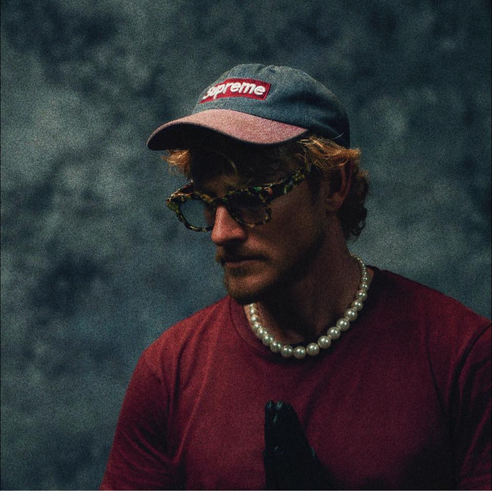 Man wearing a Supreme cap, patterned glasses, pearl necklace, maroon shirt, and black gloves against a dark textured background.