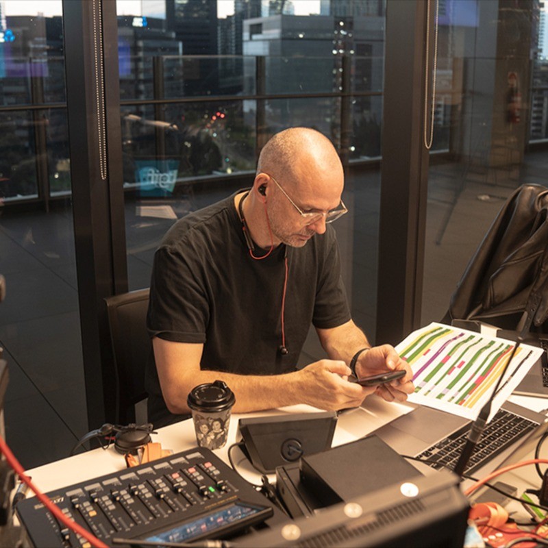 Man wearing earphones and glasses, sitting at a desk with a laptop, audio equipment, coffee cup, and a colorful chart, looking at his phone.