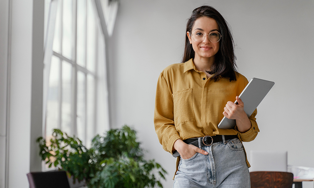 Woman with glasses in a mustard shirt holding a tablet, standing in a bright office with plants.