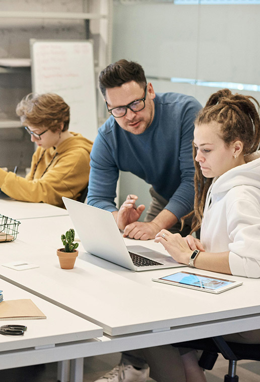 Man in blue sweater helps woman in white hoodie with laptop at a modern workspace.