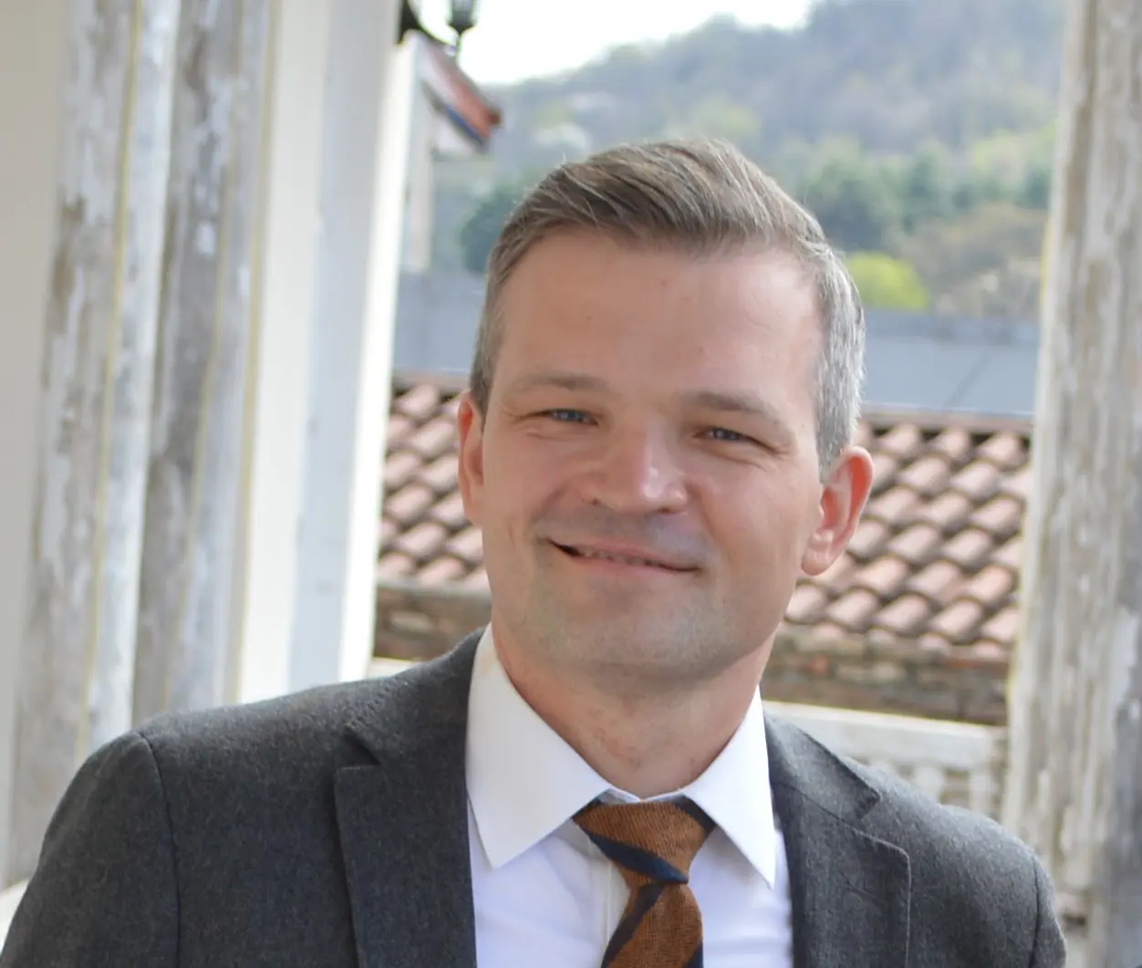 Man in a gray suit jacket representing the CEO of Detelio, white shirt, and brown tie smiling outdoors near a tiled roof.