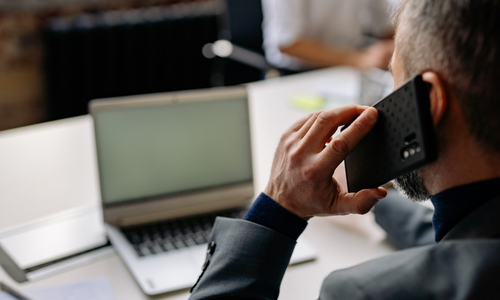 Man in a suit speaking on a smartphone with a blurred laptop on a desk in the background.