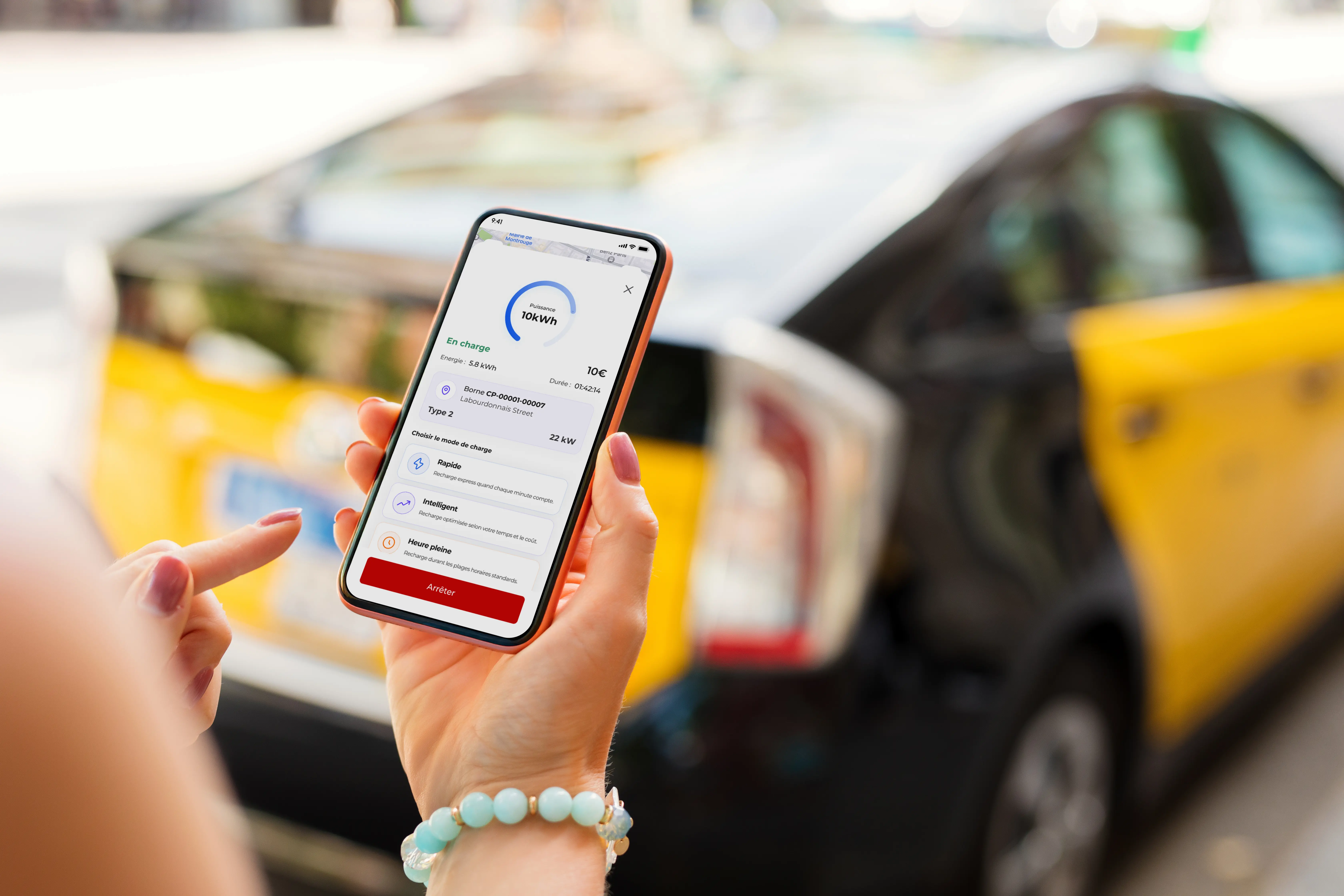 Main tenant un smartphone affichant l'application de supervision de recharge Qovoltis devant un véhicule électrique