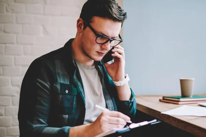 Un hombre joven, vestido casual, con lentes. Conversando por teléfono y anotando en su libreta.