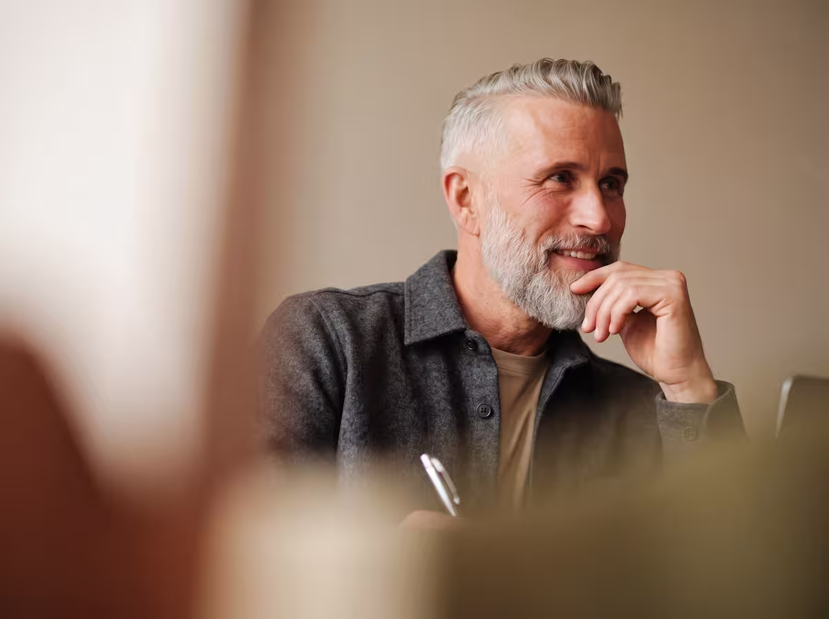 "Grey-haired bearded man in a grey wool overshirt listening attentively with his hand to his chin."