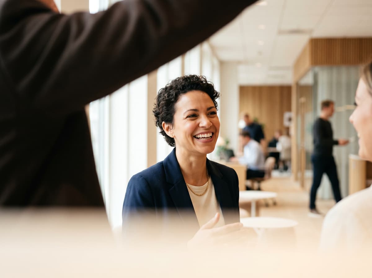 "Smiling woman in a navy blazer talking with colleagues in a bright, modern open-plan office."