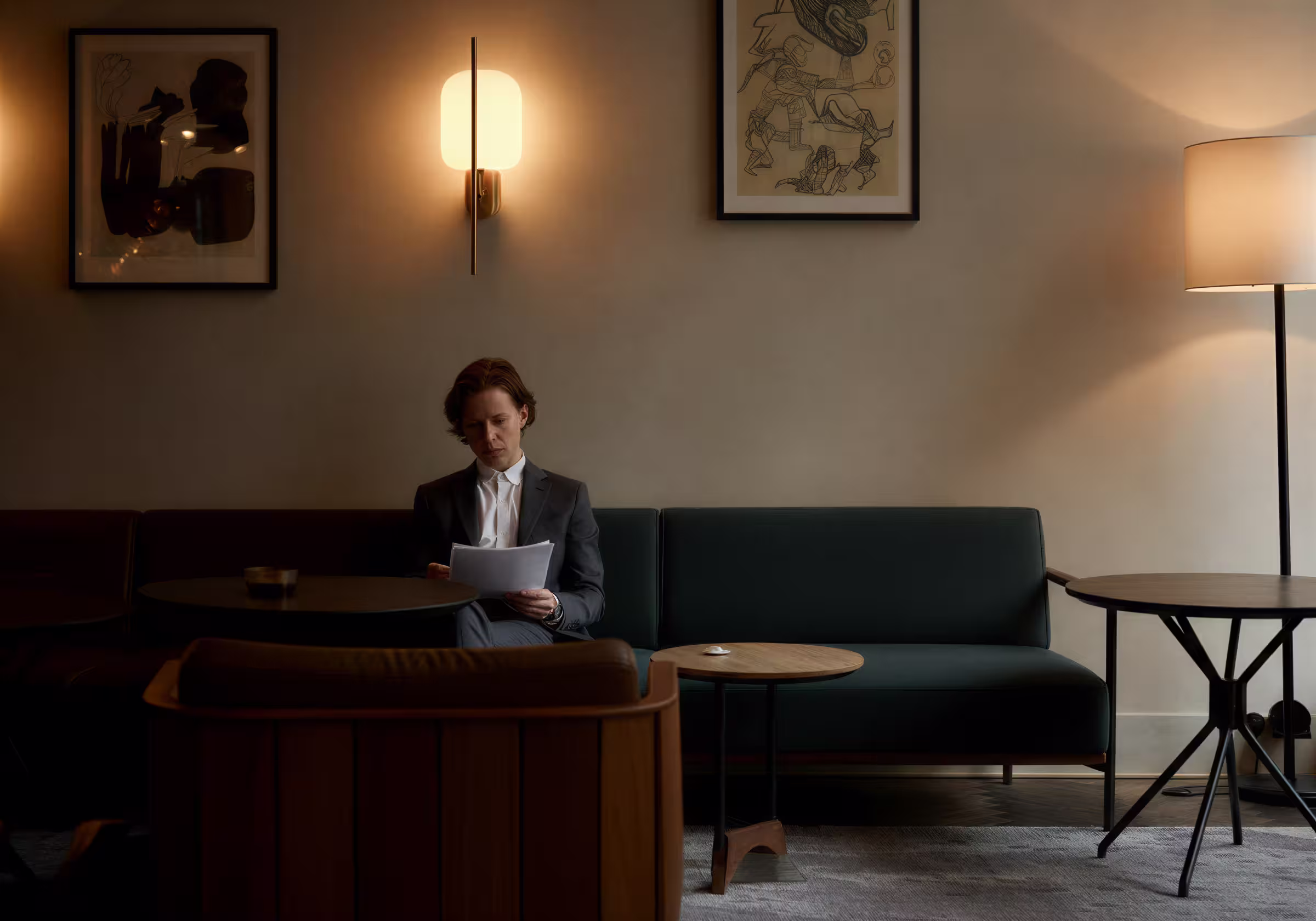 A person in a dark suit and white shirt sits on a dark green sofa, reading through some papers. The room is a stylishly decorated lounge with soft lighting, a wall-mounted lamp, a floor lamp, framed artwork on the walls, and small round wooden tables. A ca