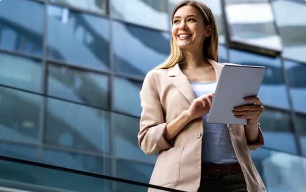 Smiling young woman in blazer holding a tablet in front of a glass building.