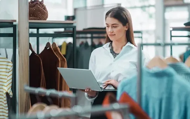 Woman using laptop while standing in clothing store near racks of hanging clothes.