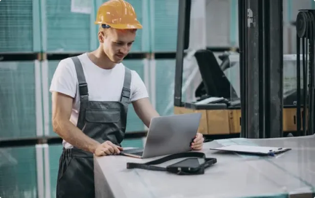 Construction worker in helmet and overalls using a laptop at a workstation.