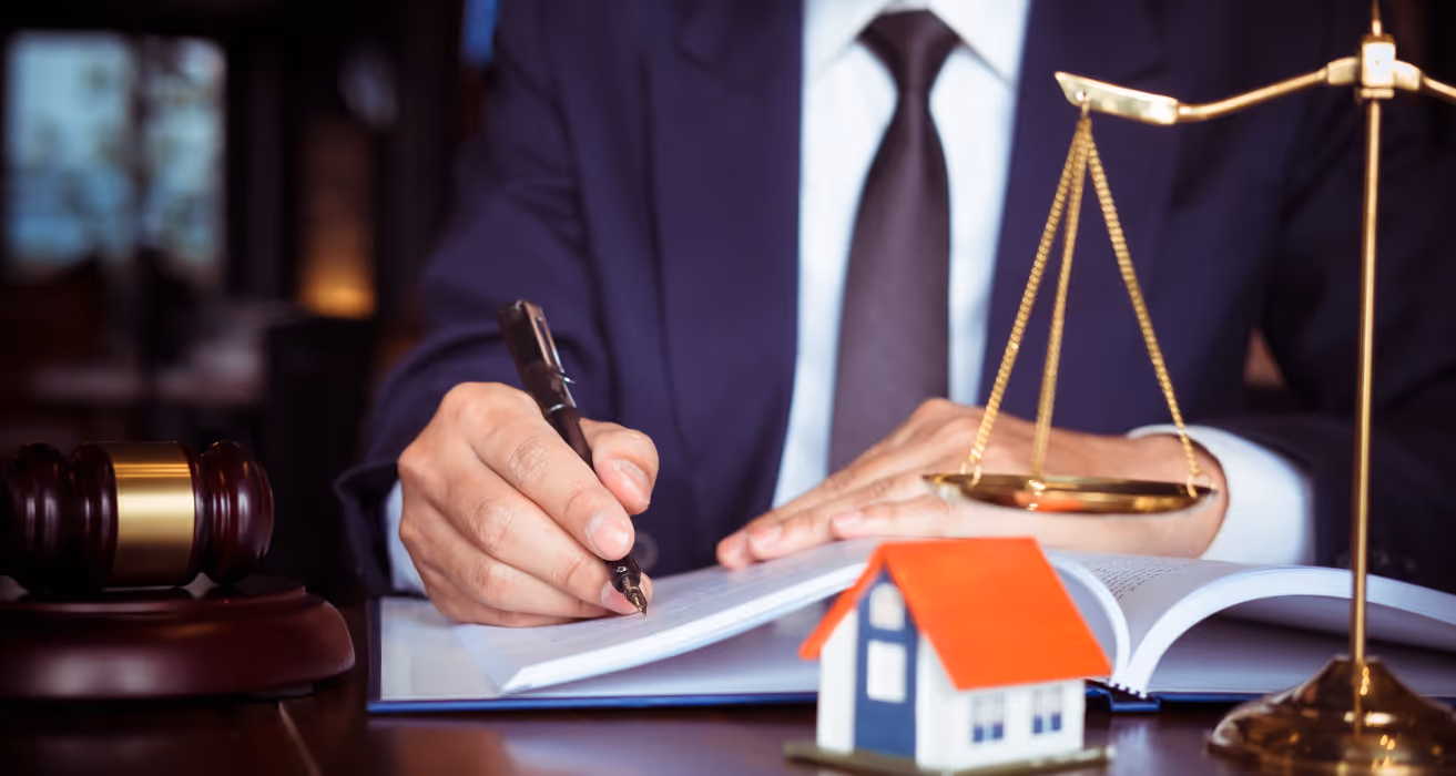 Lawyer signing legal documents with gavel, scales, and house model on desk, symbolizing real estate law and mortgage agreements.