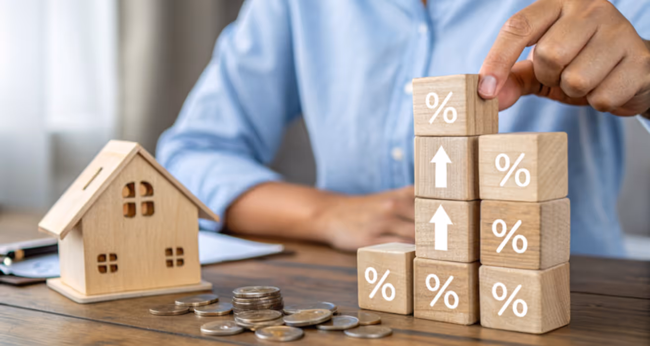 Person stacking wooden blocks with percentage signs and arrows beside a house model and coins, symbolizing rising interest rates.