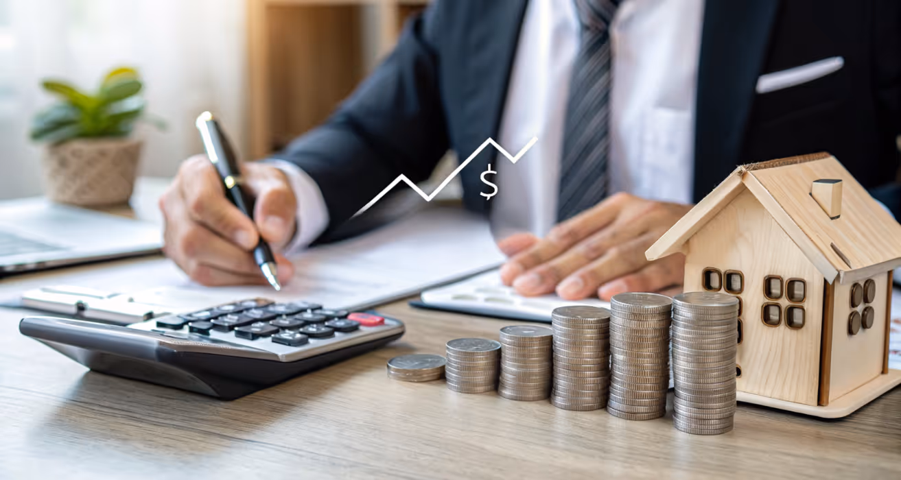 Businessperson calculating real estate investment returns with stacked coins, house model, and financial growth chart on desk.