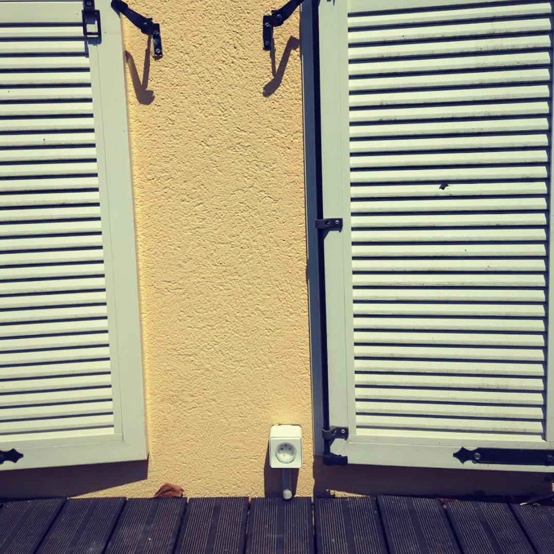 Prise extérieure étanche installée sur un mur de terrasse, avec volets en arrière-plan