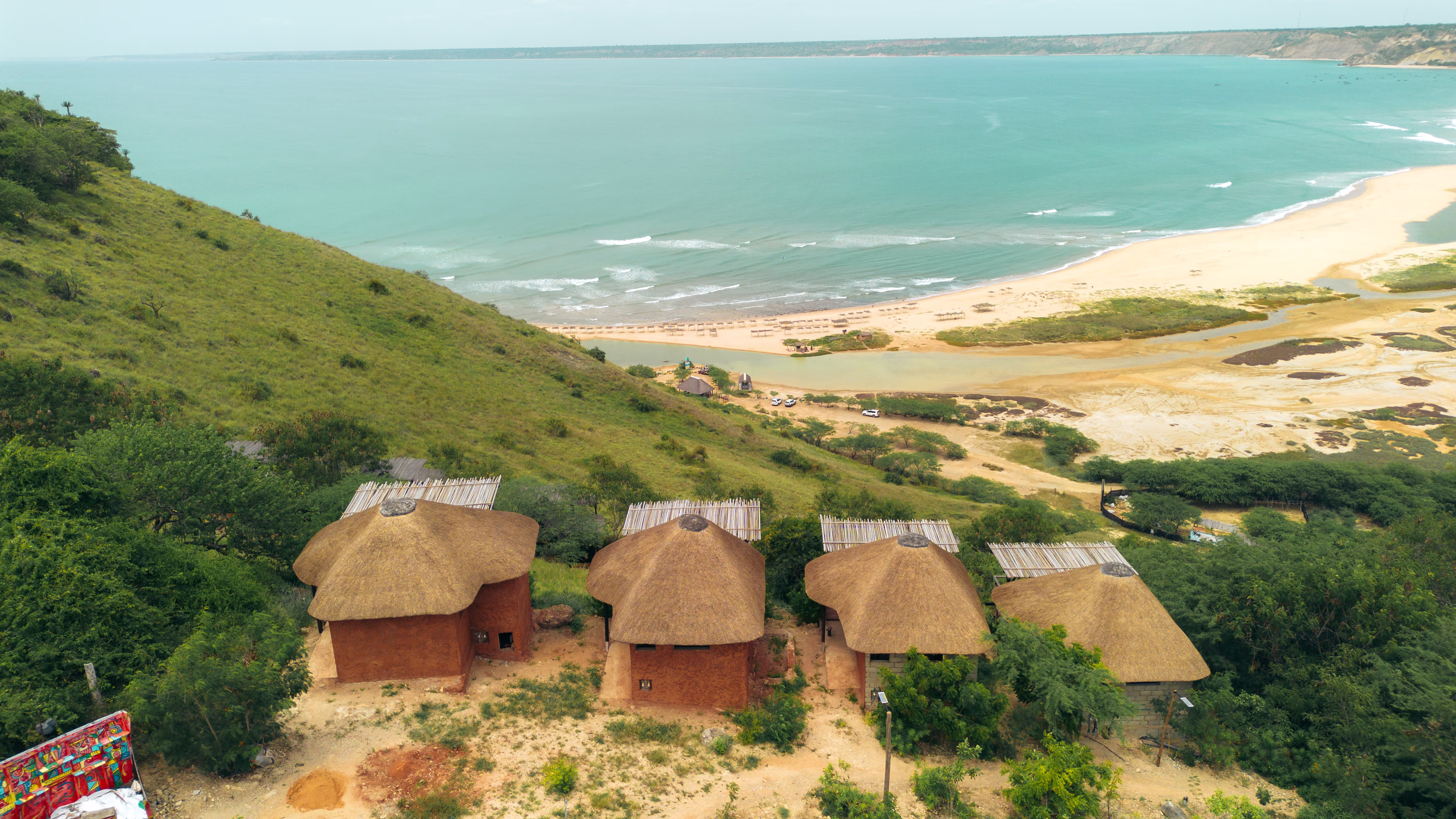 Vier traditionelle Strohhütten mit Reetdächern auf einem grünen Hügel über dem sandigen Strand von Cabo Ledo und dem weiten Atlantik im Hintergrund.