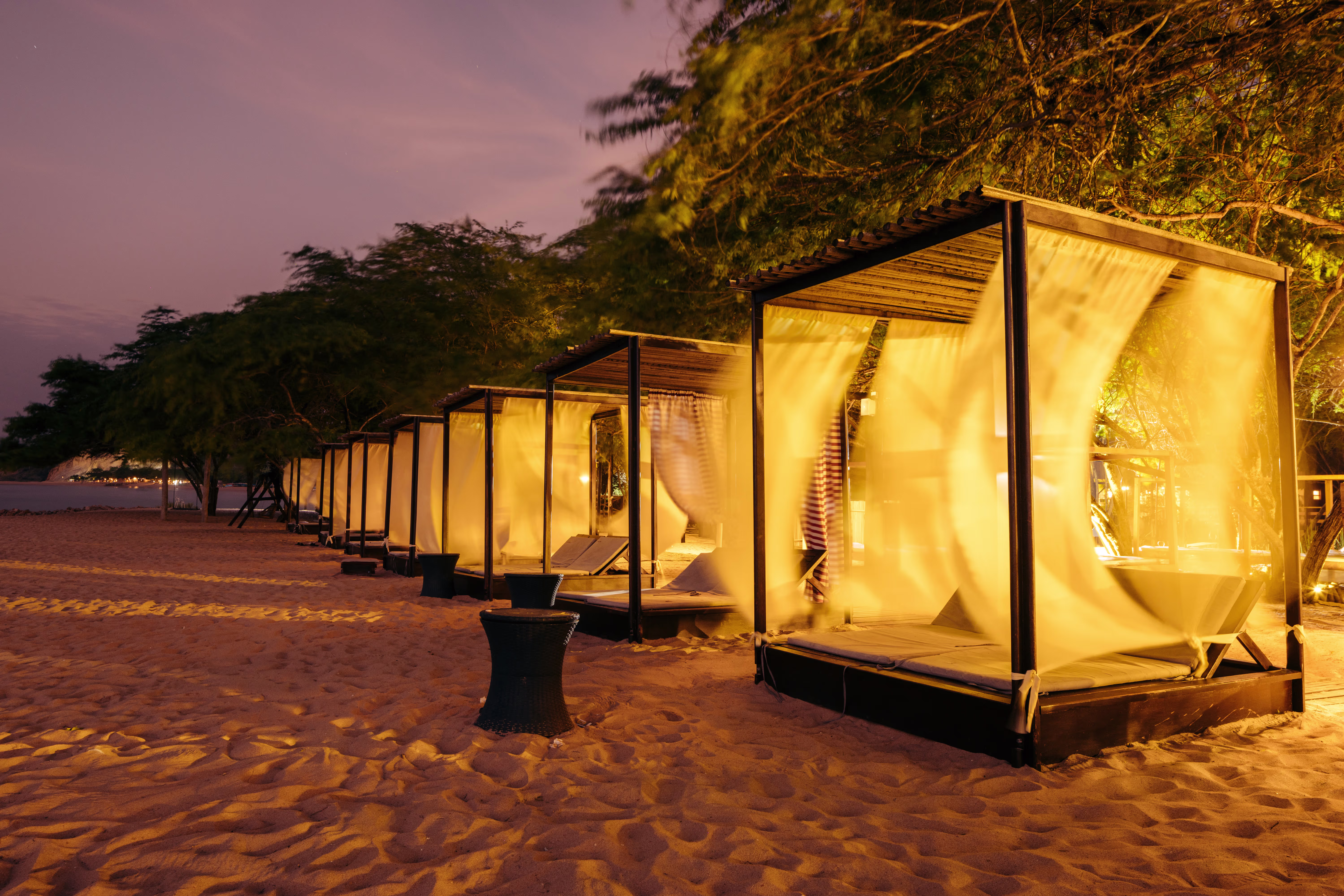 Reihe eleganter Beachbeds mit leichten Vorhängen auf dem Sandstrand bei Dämmerung, umrahmt von Küstenbäumen.