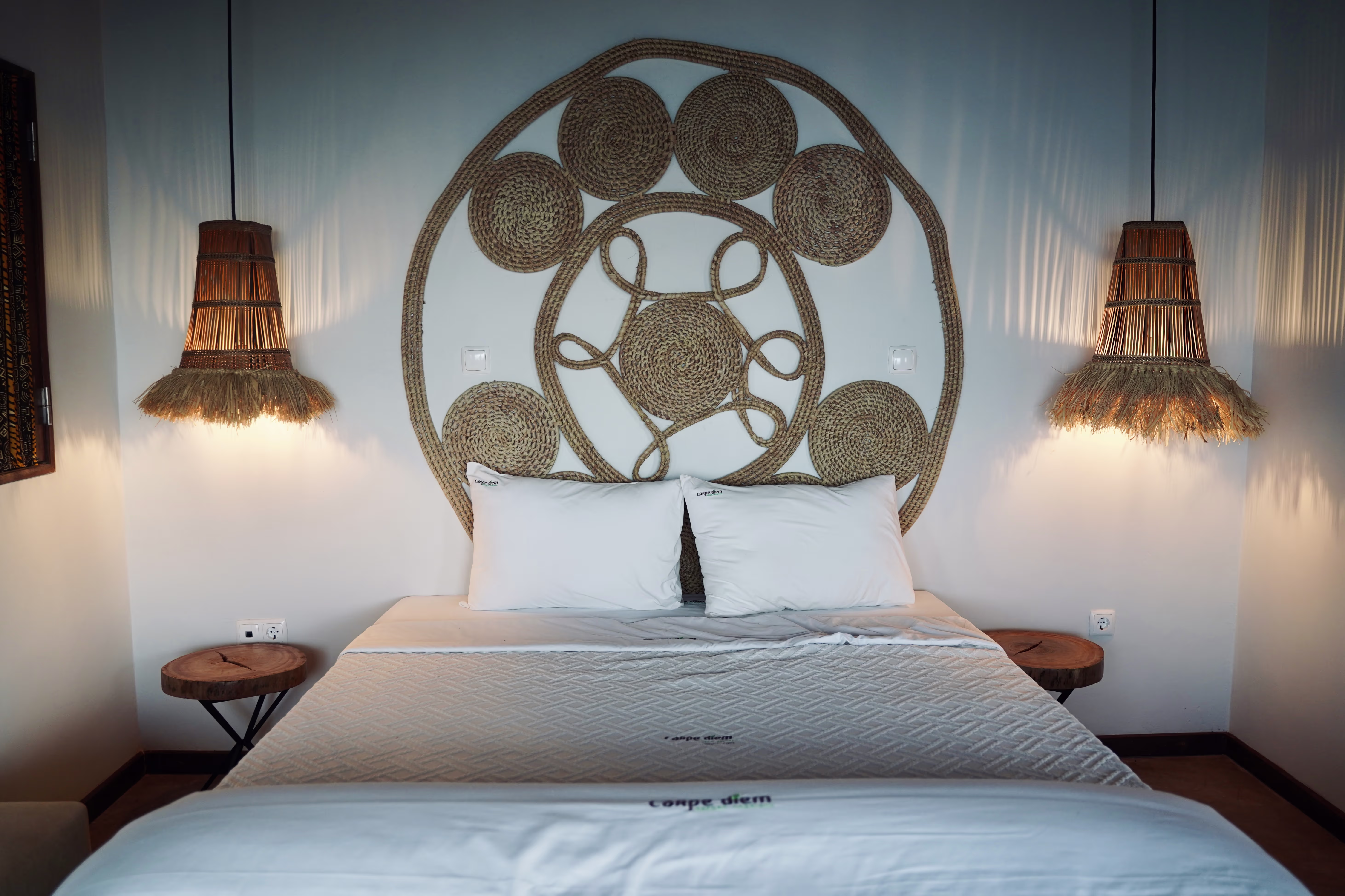 Elegantes Doppelbett mit weißer Bettwäsche vor einer runden Wanddekoration aus geflochtenem Seegras und seitlichen Lampen mit Fransen.