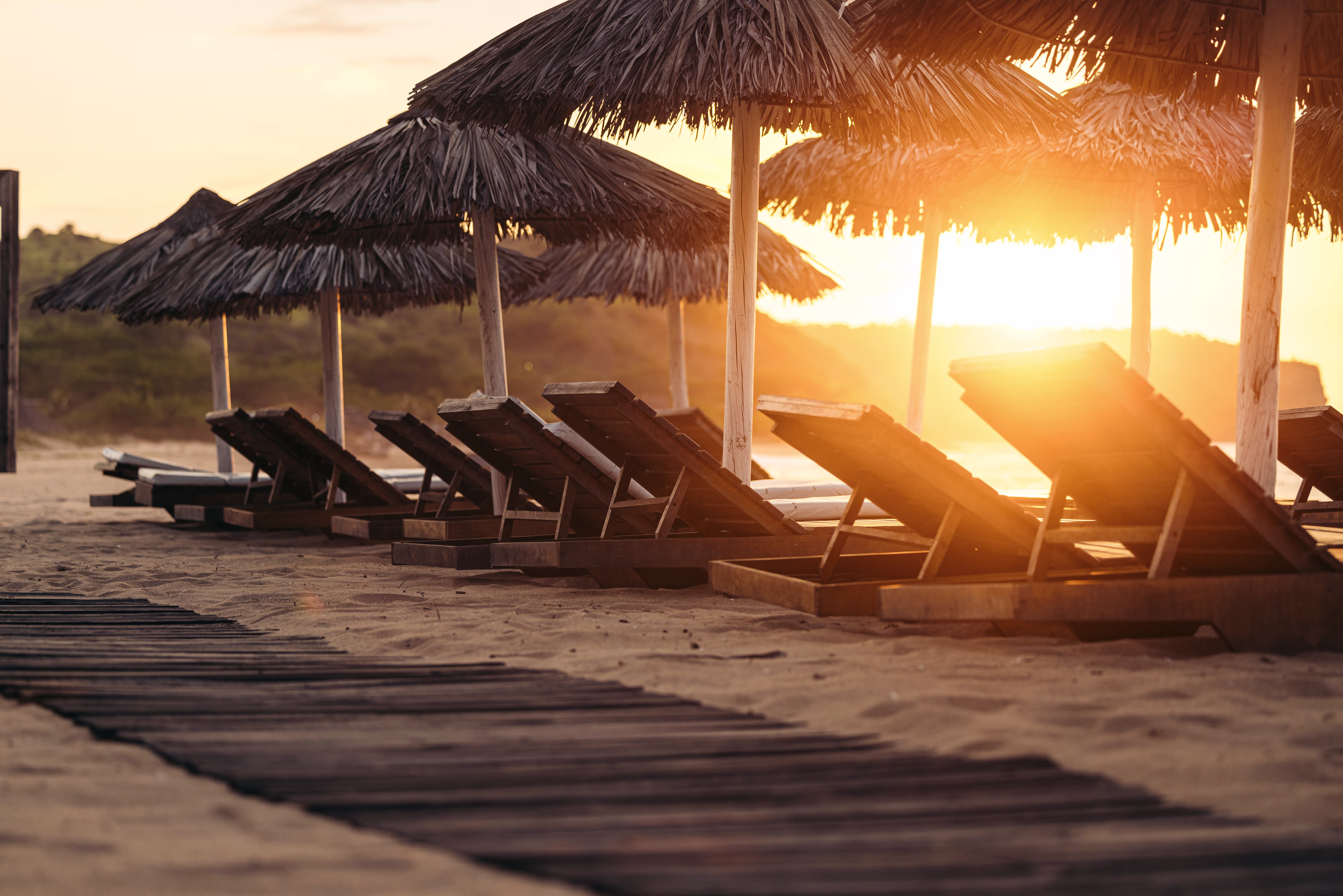 Reihe rustikaler Holzliegen unter Strohdächern am Sandstrand bei Sonnenuntergang, mit warmem Abendlicht.“
