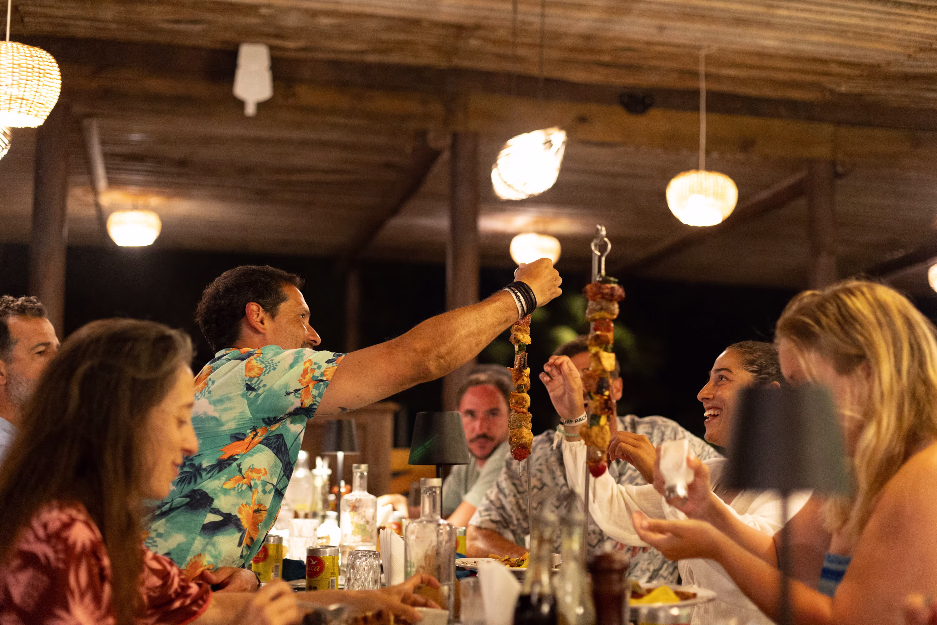 Gäste im Carpe Diem Resort sitzen an einem langen Holztisch und stoßen mit gegrillten Spießen und Drinks in einer offenen Strandbar an