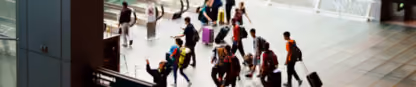 Travelers with luggage walk through busy airport terminal, heading to escalators amidst directional signs and bustling activity.