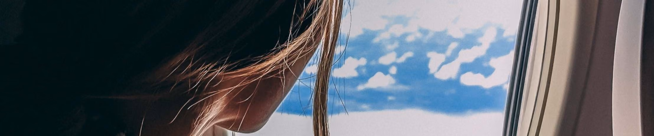 Woman gazing out airplane window at clouds and blue sky, experiencing flight travel and aerial view from cabin seat.