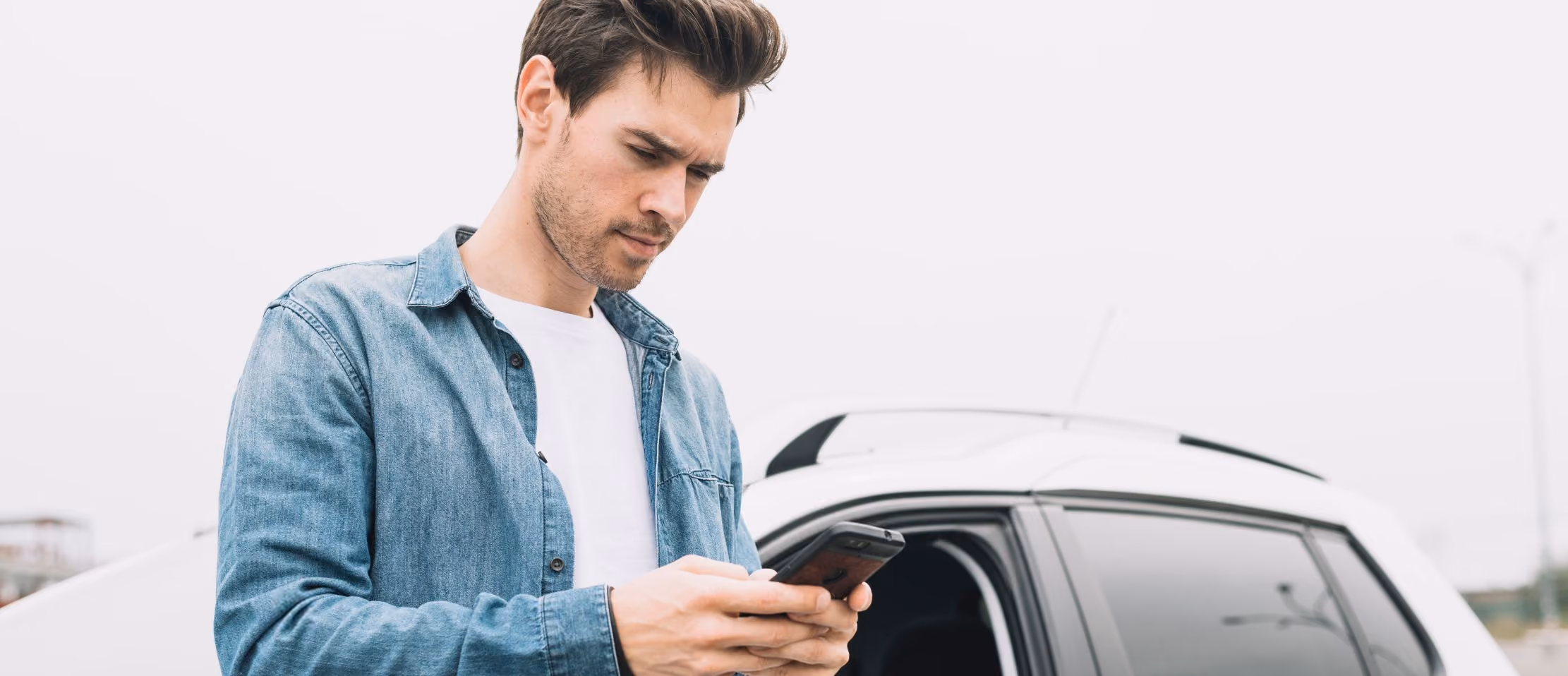 Man using smartphone near car, wearing denim jacket; likely searching or messaging while standing outdoors.