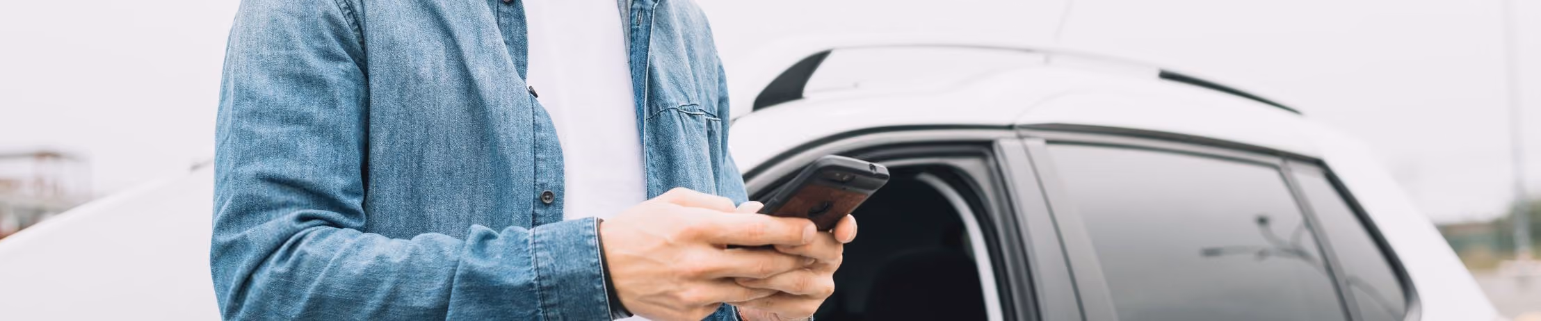 Man using smartphone near car, wearing denim jacket; likely searching or messaging while standing outdoors.
