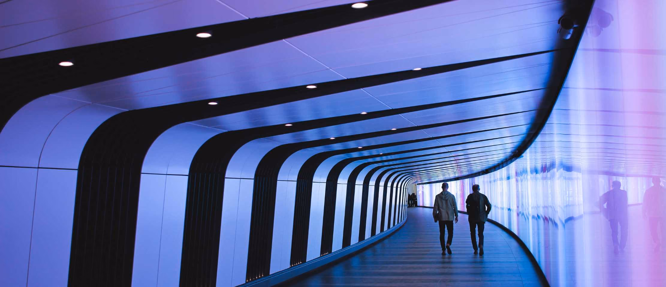 Futuristic tunnel with purple lights, two people walking, modern architecture.