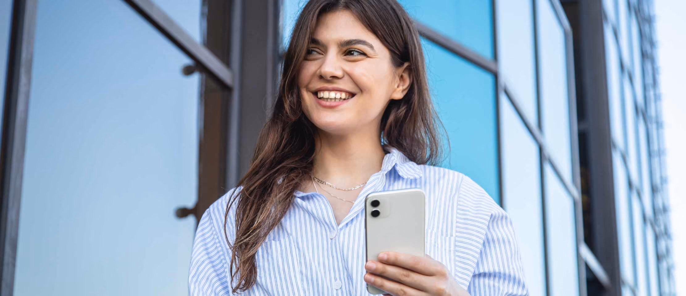 Smiling woman holding smartphone near modern glass building, ideal for tech and lifestyle content.