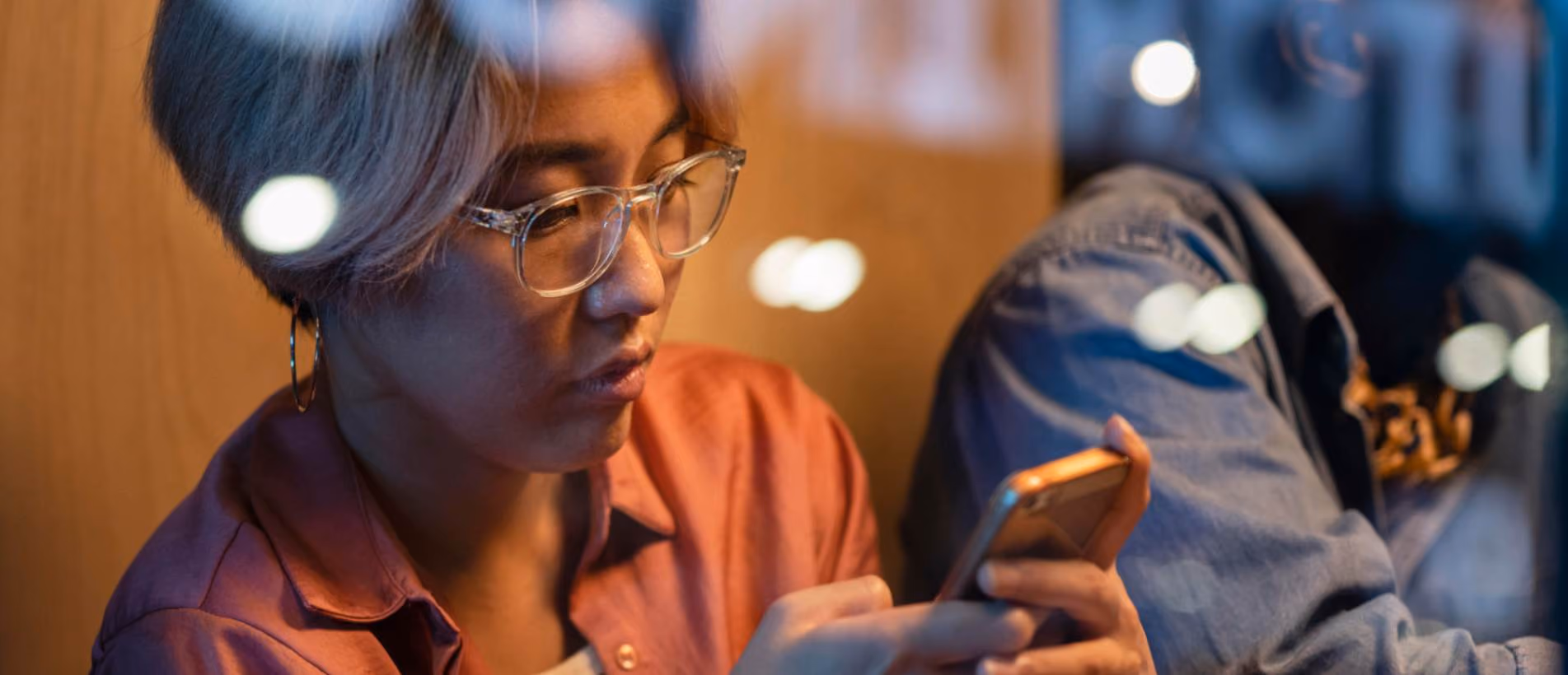 Person focused on smartphone screen in dimly lit setting, wearing glasses and casual attire, reflecting mobile technology usage.