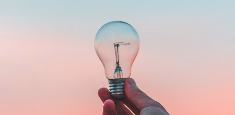 Hand holding lightbulb against pastel sky, symbolizing creativity and innovation.