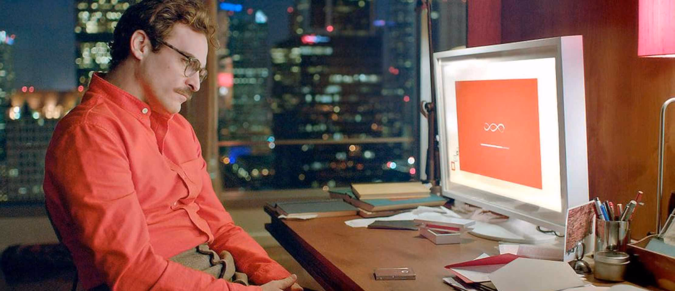 Man in red shirt at a desk with computer screen, overlooking cityscape at night. Perfect for tech and lifestyle articles.