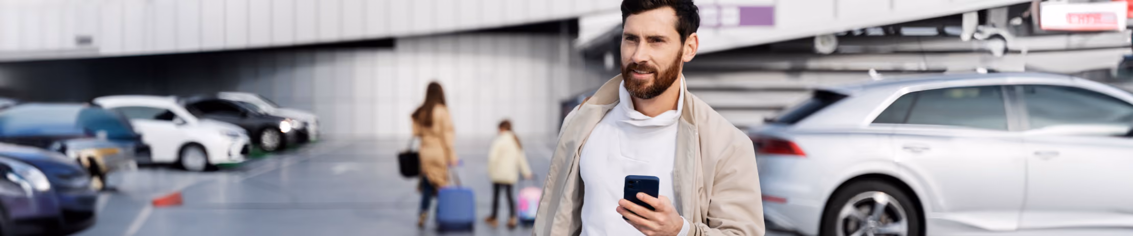 Man walking in parking lot, pulling suitcase and using smartphone, with cars and family in background, capturing travel convenience.