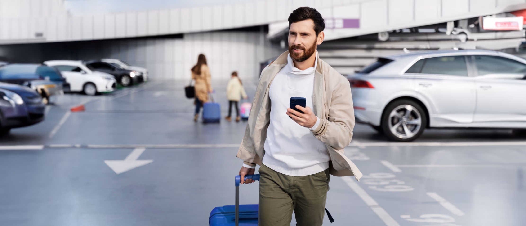 Man walking in parking lot, pulling suitcase and using smartphone, with cars and family in background, capturing travel convenience.