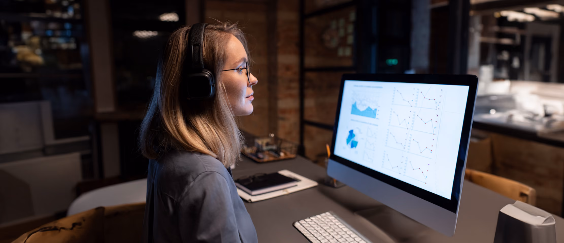 Woman analyzing graphs on a computer, wearing headphones, focused on data visualization in a modern office setting.