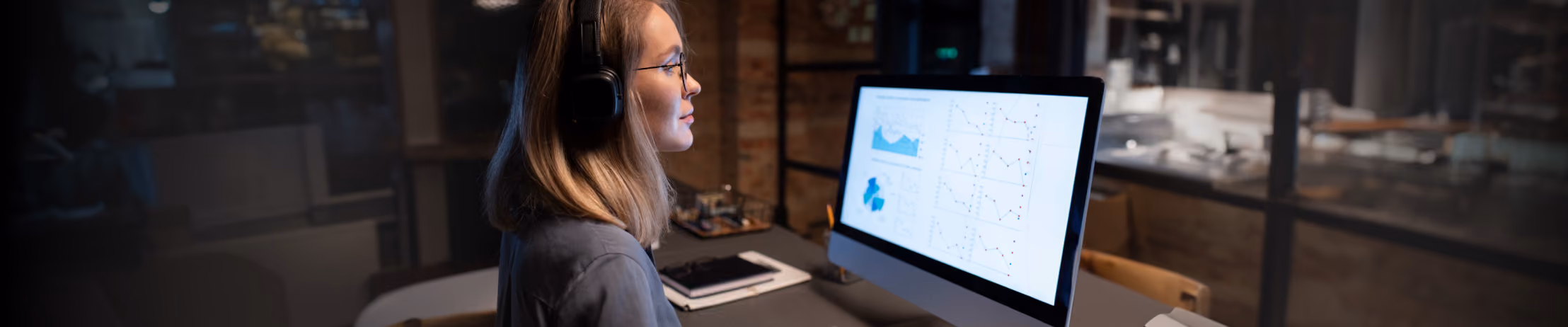 Woman analyzing graphs on a computer, wearing headphones, focused on data visualization in a modern office setting.