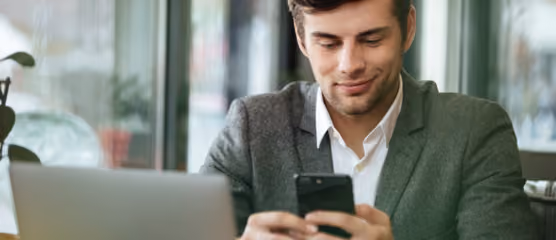 Man in a suit smiling at smartphone in cafe, working on laptop, highlighting business communication and remote work.