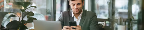 Man in a suit smiling at smartphone in cafe, working on laptop, highlighting business communication and remote work.
