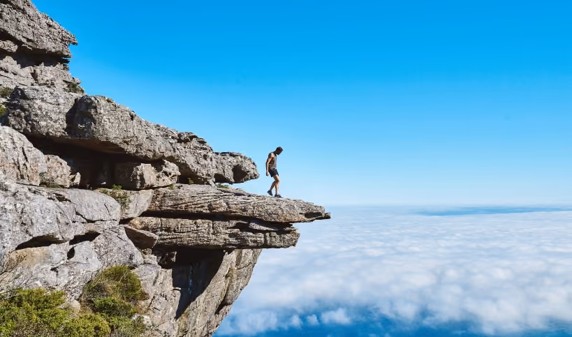 Person standing on cliff edge above clouds, symbolizing risk-taking and adventure.