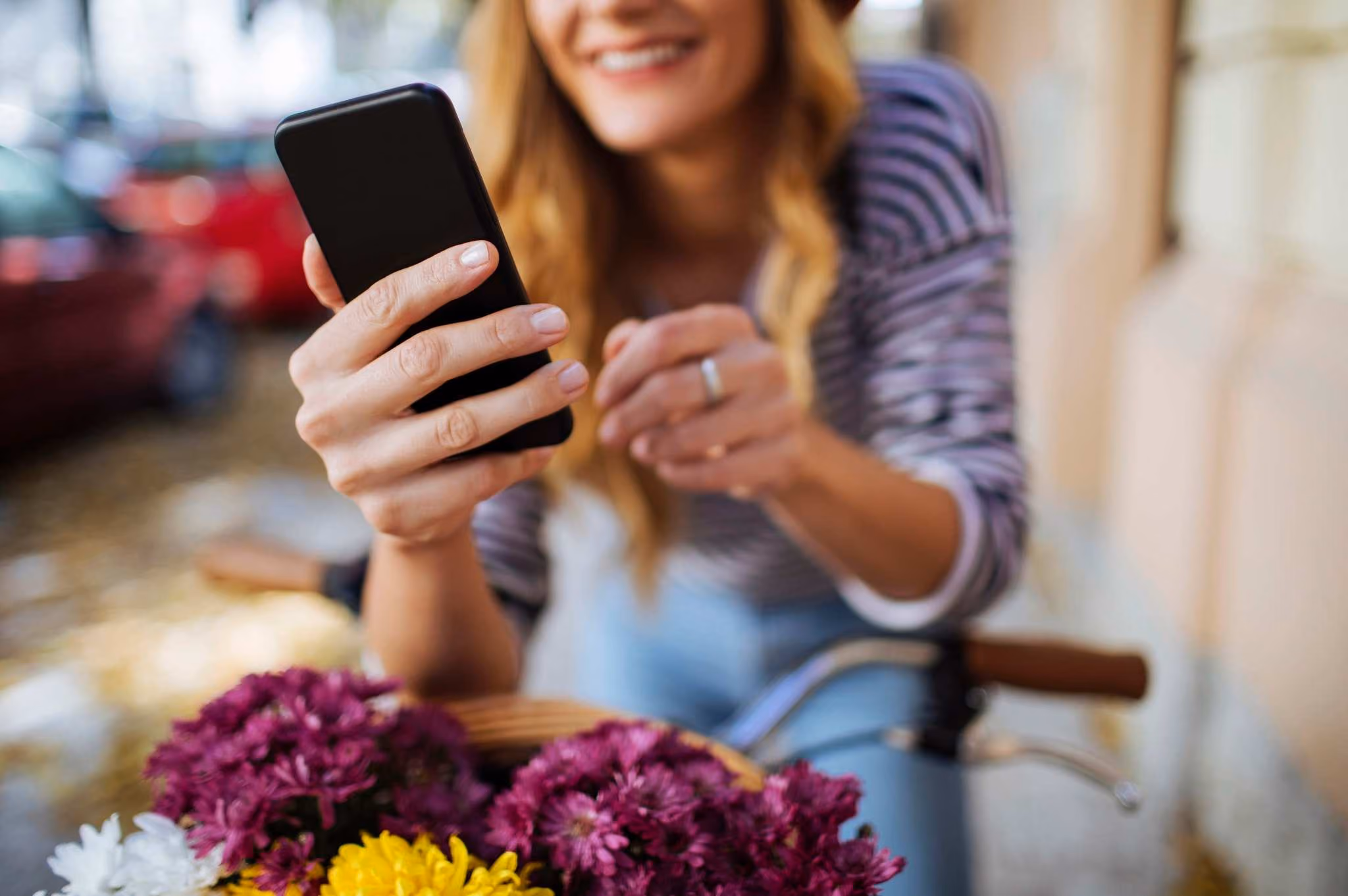 Woman on bike holding smartphone and smiling, with a basket of colorful flowers, outdoor urban setting.