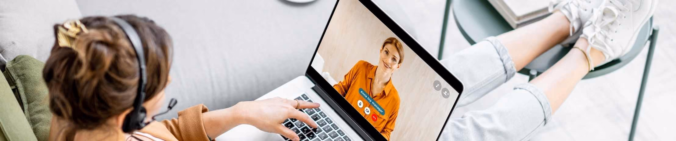 Person in headset on video call using laptop, reclining on sofa with feet on table, coffee cup nearby, home office setting.