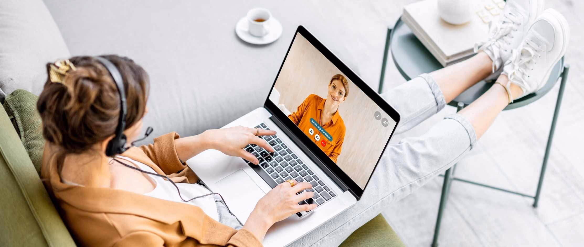 Person in headset on video call using laptop, reclining on sofa with feet on table, coffee cup nearby, home office setting.