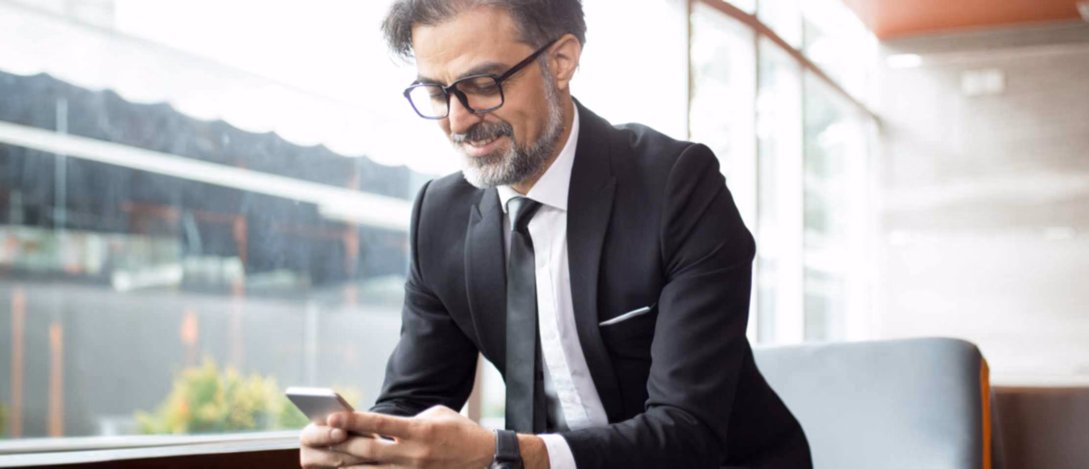 Businessman in suit sitting by window, using smartphone, smiling.