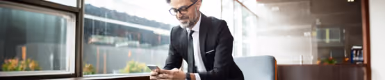 Businessman in suit sitting by window, using smartphone, smiling.
