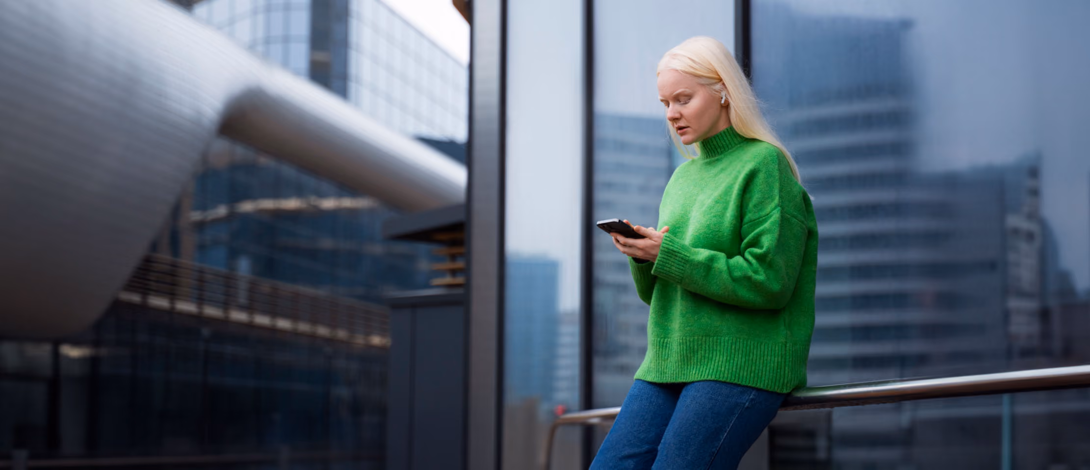 Woman in green sweater using smartphone with earbuds outside modern city building, focusing on communication and technology.