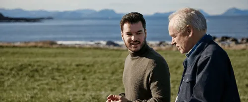 Bildet viser to menn som går og prater på en gresslette ved havet, hvor den yngre mannen har på seg en grønn genser og den eldre mannen en blå jakke. Bak dem kan man se fjorder og øyer i det fjerne.