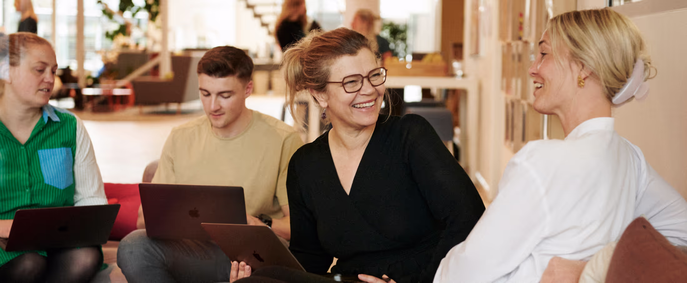 Bildet viser en gruppe mennesker som smilende diskuterer i en uformell sittegruppe, hvor to av dem holder bærbare datamaskiner. En kvinne i sorte briller ler sammen med en kvinne i hvit topp, omgitt av et moderne kontormiljø.